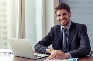 Portrait of handsome young businessman in formal suit using a laptop, looking at camera and smiling while working in office